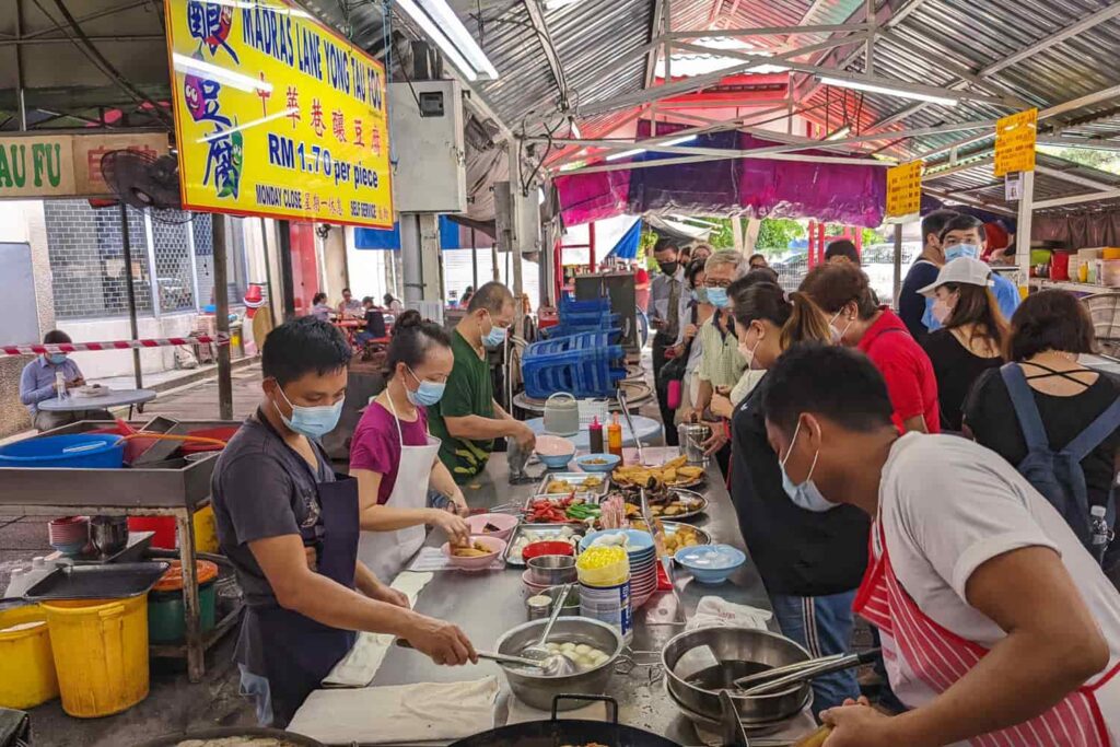 Queue of people along a table of yong tau foo with hawkers cooking and selling near Petaling Street Chinatown