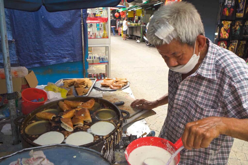 Man making mini apom balik pancakes standing at hawker cart with batter