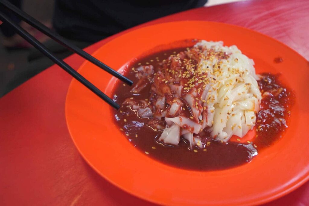 Red plate with clear chee cheong fun noodles covered in red sweet sauce and sesame seeds