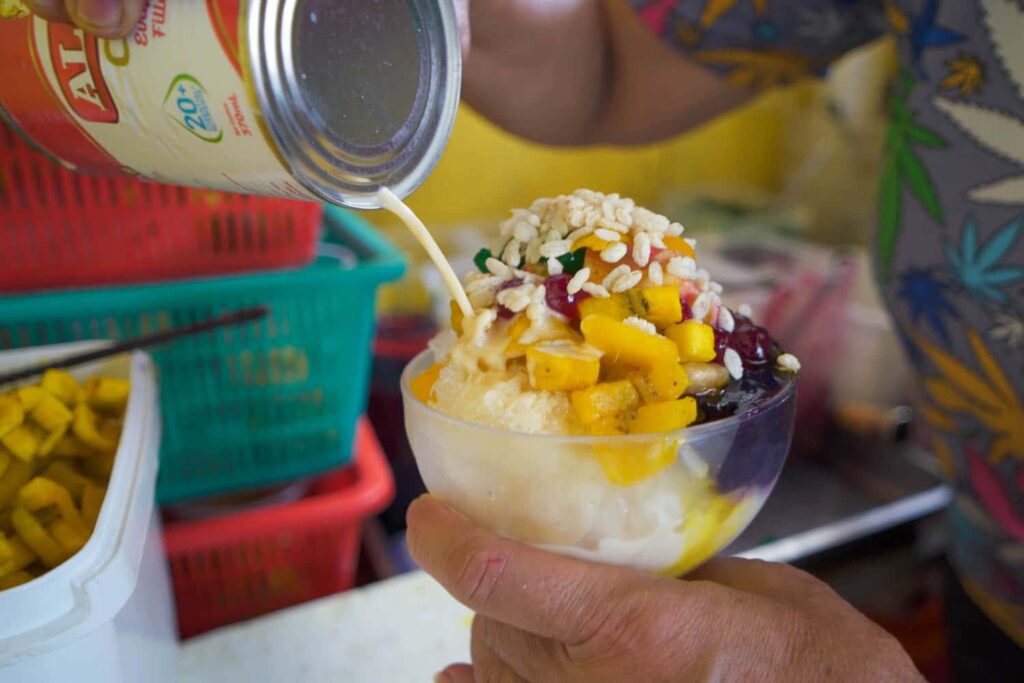 Evaporated milk pouring onto bowl of halo-halo, bananas, ube jam, shaved ice