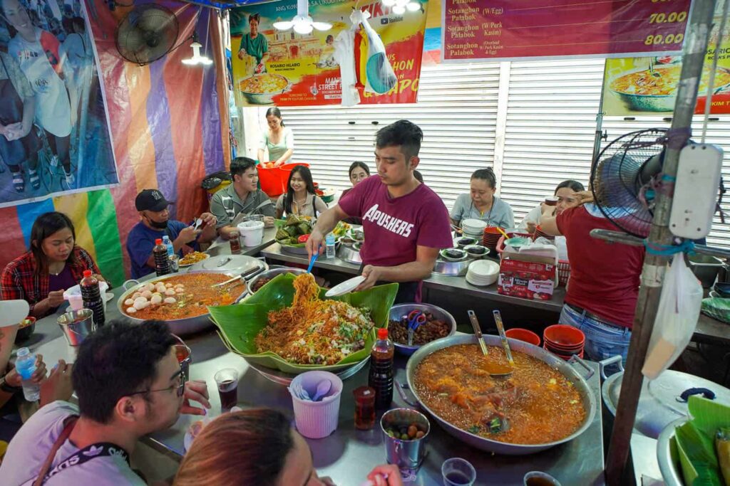 Rosa's Pancitan Famous Sotanghon Quiapo Market man serving noodles with three large woks customers seated around
