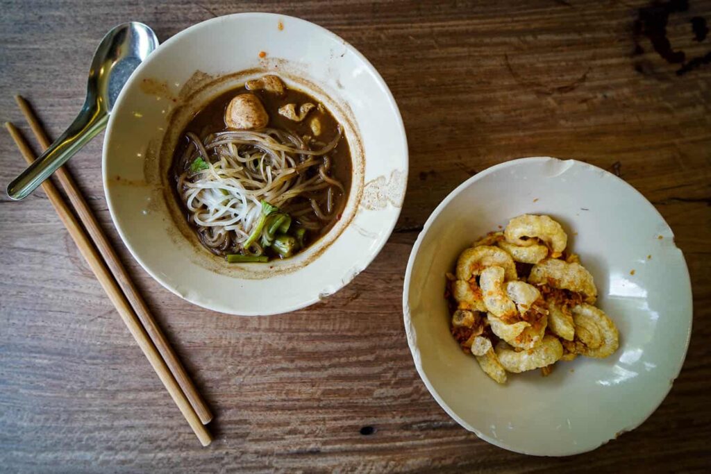 Two bowls on wooden table, boat noodles and crispy pork skin