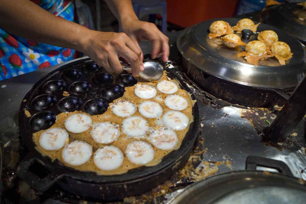 Person scooping out khanom krok thai street food with spoon out of iron grill