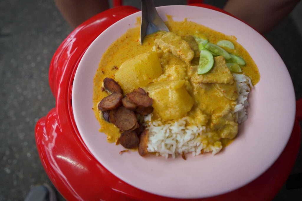 pink plate of yellow thai curry on bed of rice sitting on red plastic stool on street