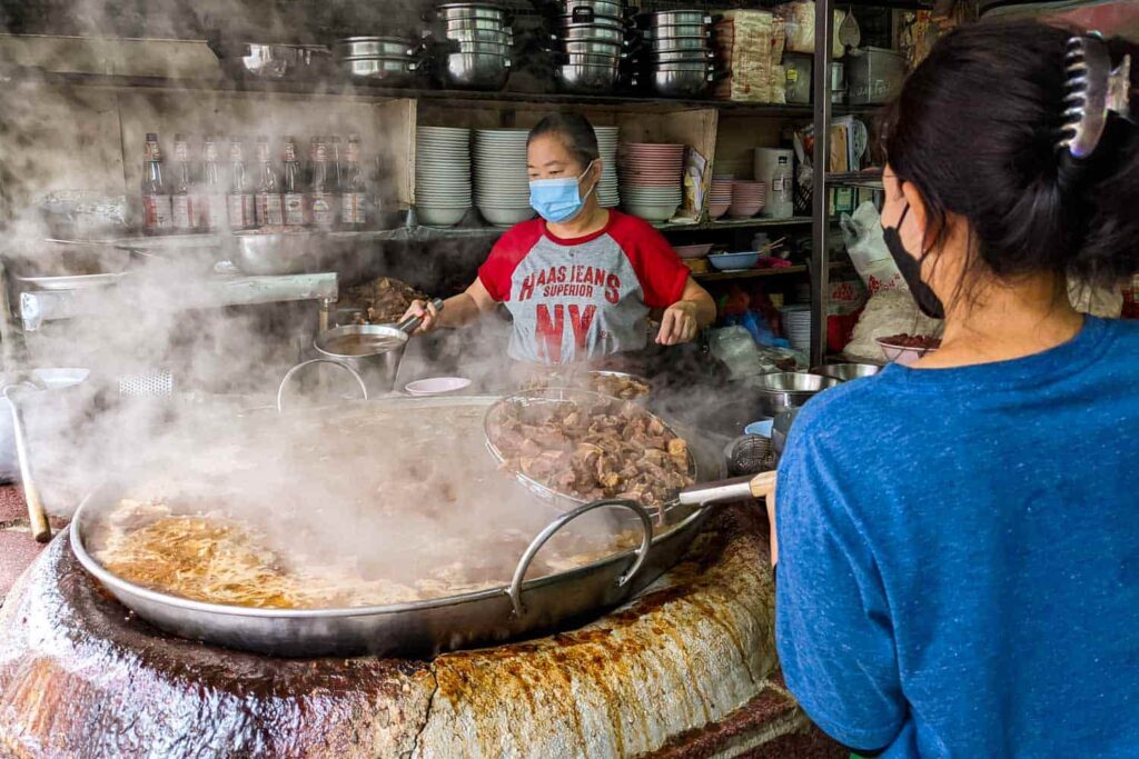 Large vat of beef stew being stirred by two women in thai street food shop