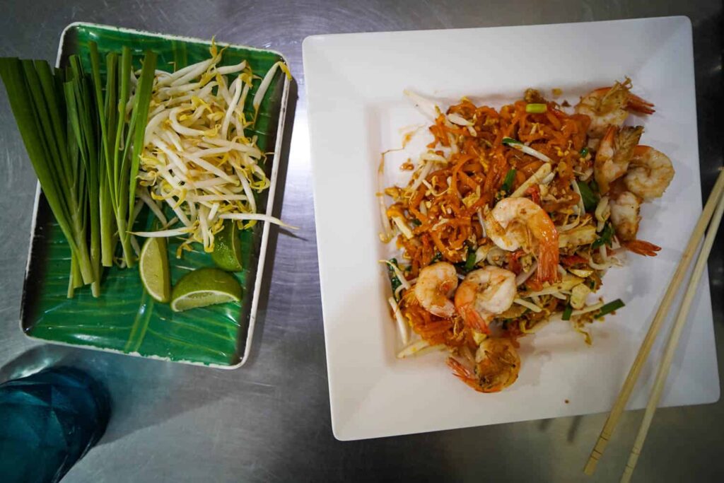 Plate of shrimp pad thai on metal table with fresh spring onion and bean sprouts