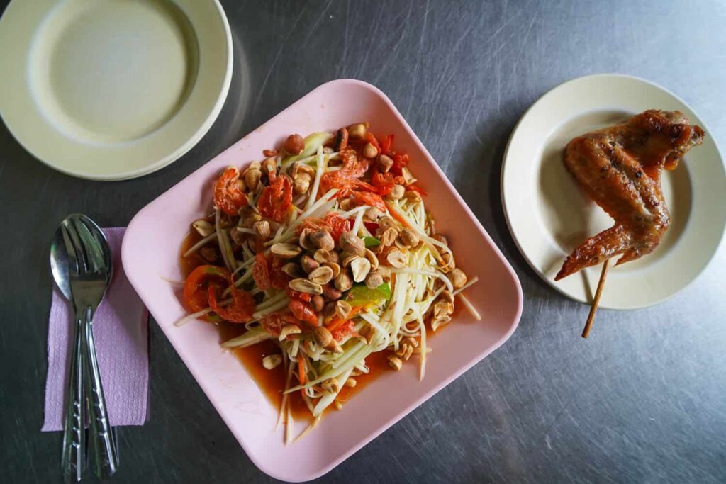 Plate of papaya salad with peanuts and dried shrimp with chicken wing on metal table