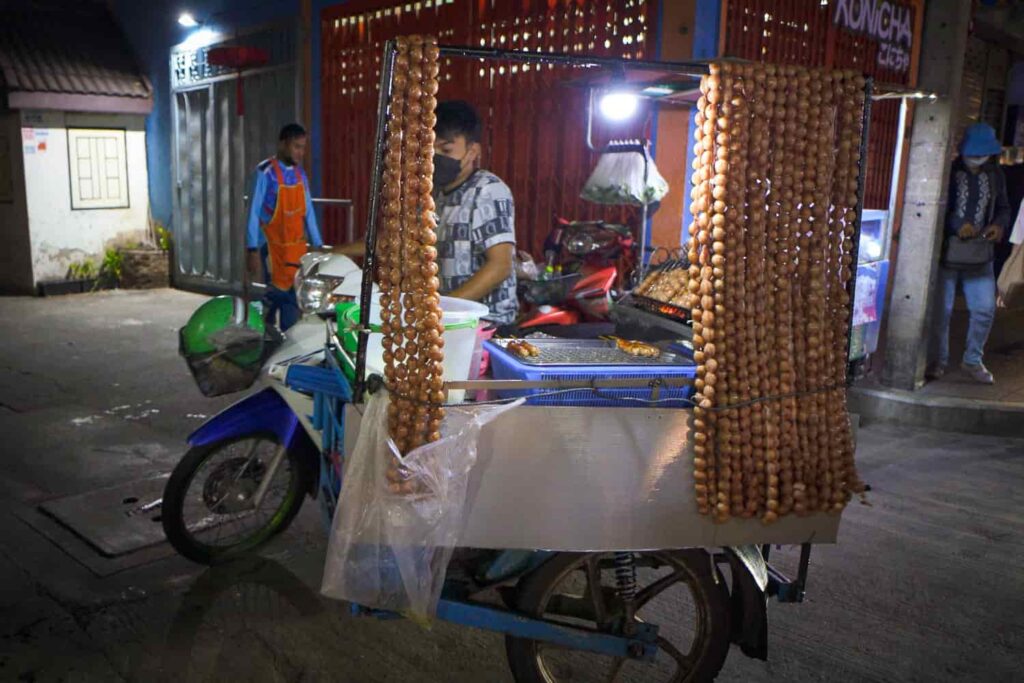 Man on motorbike attached to thai street food cart selling dozens of strings of sai krok