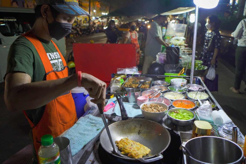 Thai man cooking thai omelette in wok with bowls of vegetables on thai street cart