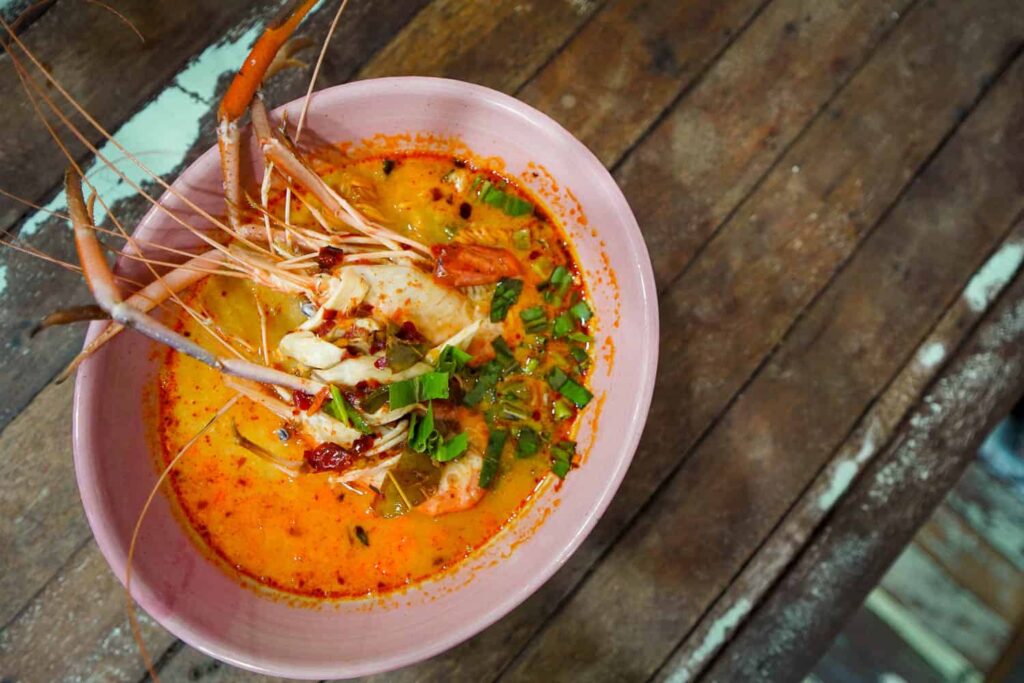 Bowl of orange tom yum soup with large prawn in bowl on wooden table