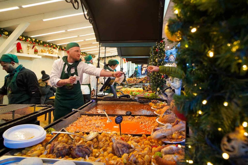 Several trays of local Hungarian food being sold by people in green with a Christmas tree in the foreground.