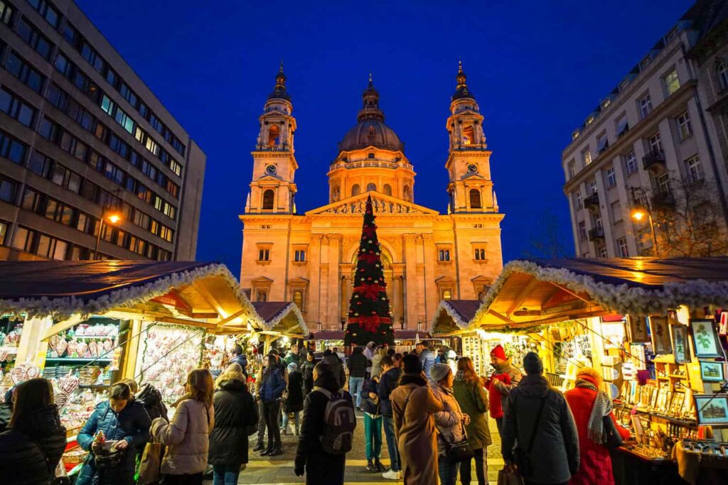 Nighttime scene of St. Stephen's Basilica, with two rows of wooden huts selling local artisan crafts to shoppers in the centre.