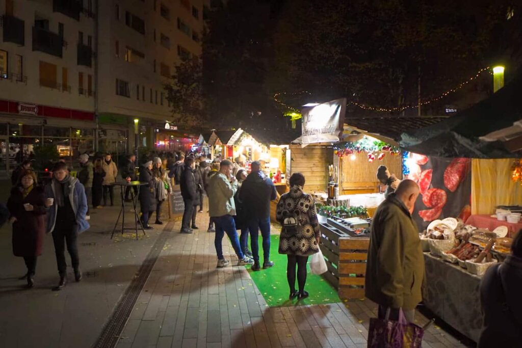 People standing outside of wooden huts which are selling sausage and Hungarian foods.