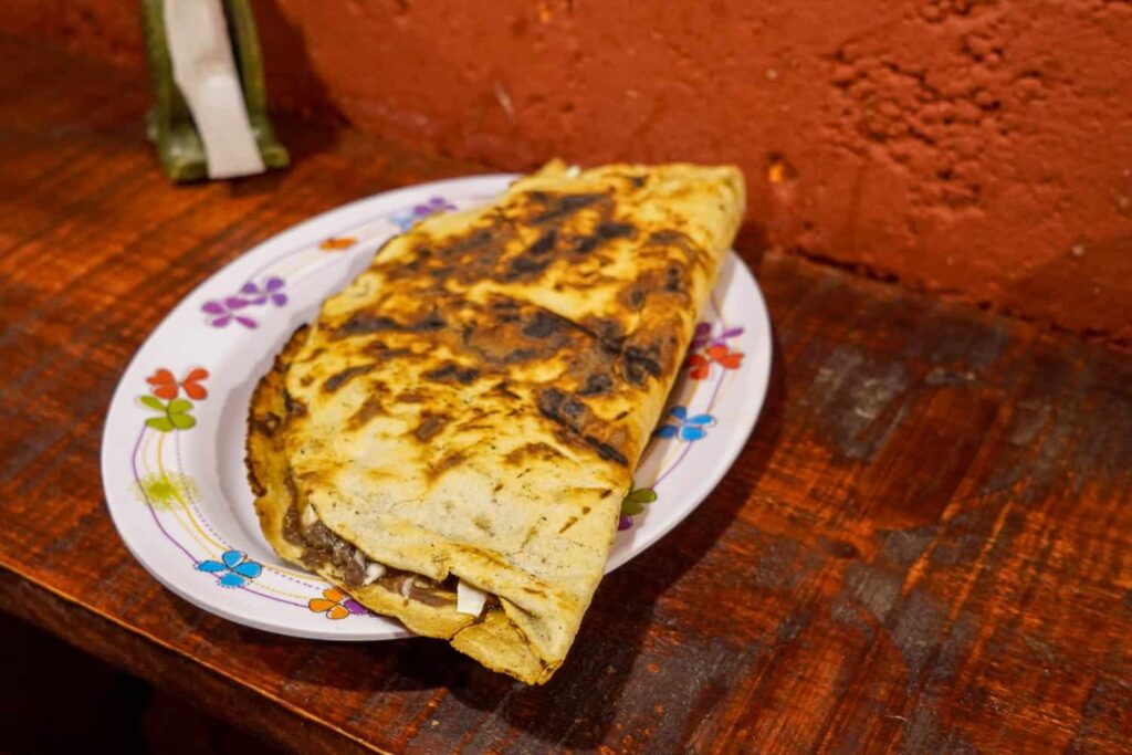 Crispy grilled tlayuda with quesillo and meat on a plate placed on a red bench at Tlayudas Don Ramón, known for the best tlayudas in Oaxaca.