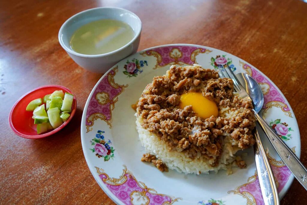 Plate of Nasi Tim chicken rice with minced chicken and raw egg yolk, served with soup and pickled veggies.