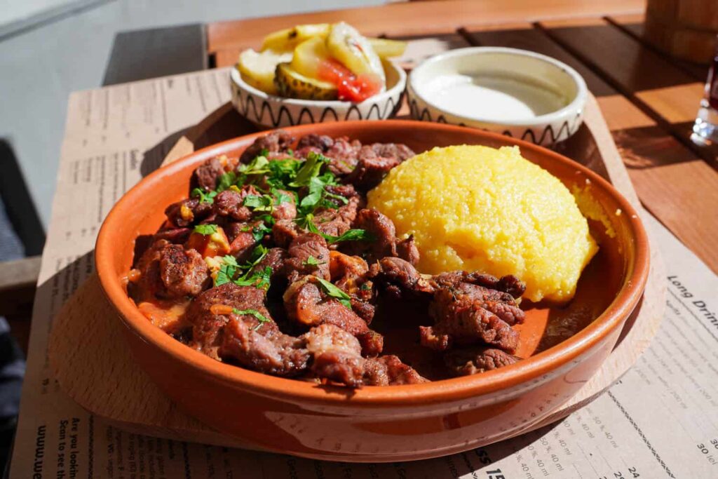 Transylvanian pork stew with mamaliga polenta in a clay bowl on a rustic wooden table in the sun
