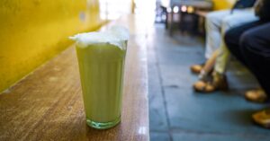 Cold and delicious glass of the best lassi in Mumbai, thick and white with malai on top, sitting on a brown bench with yellow wall behind in small lassi shop