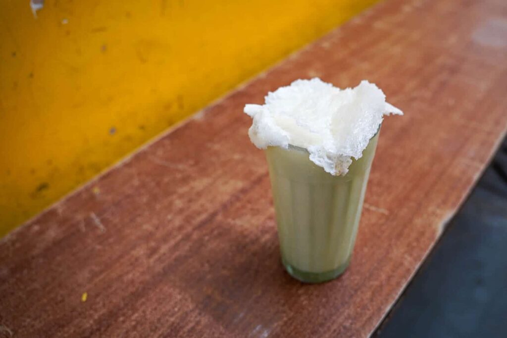 A cold glass of the best lassi in Mumbai with malai on top sitting on brown bench with yellow wall behind